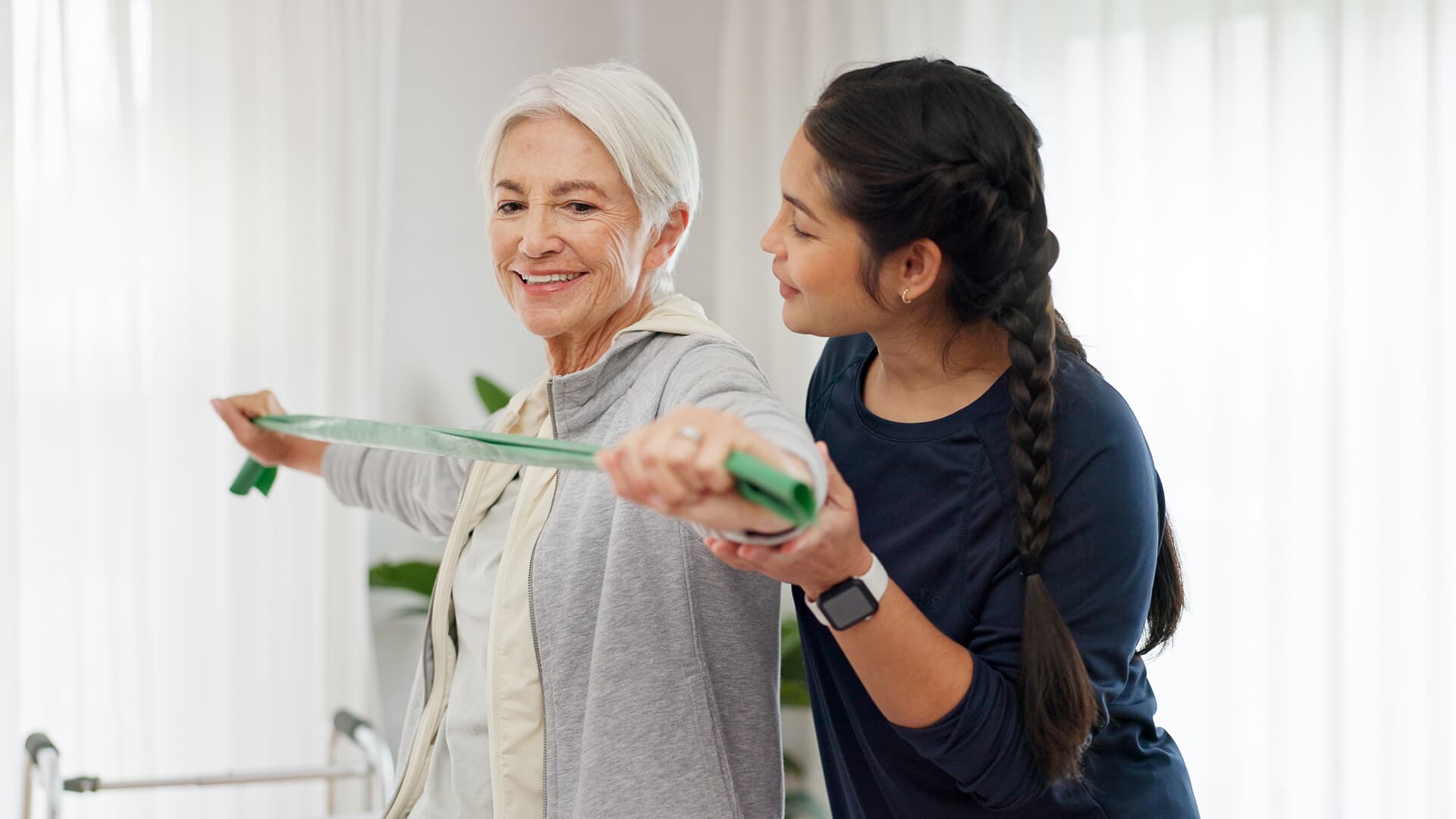 Ältere Frau trainiert gemeinsam mit einer Pflegekraft mit einem elastischen Trainingsband