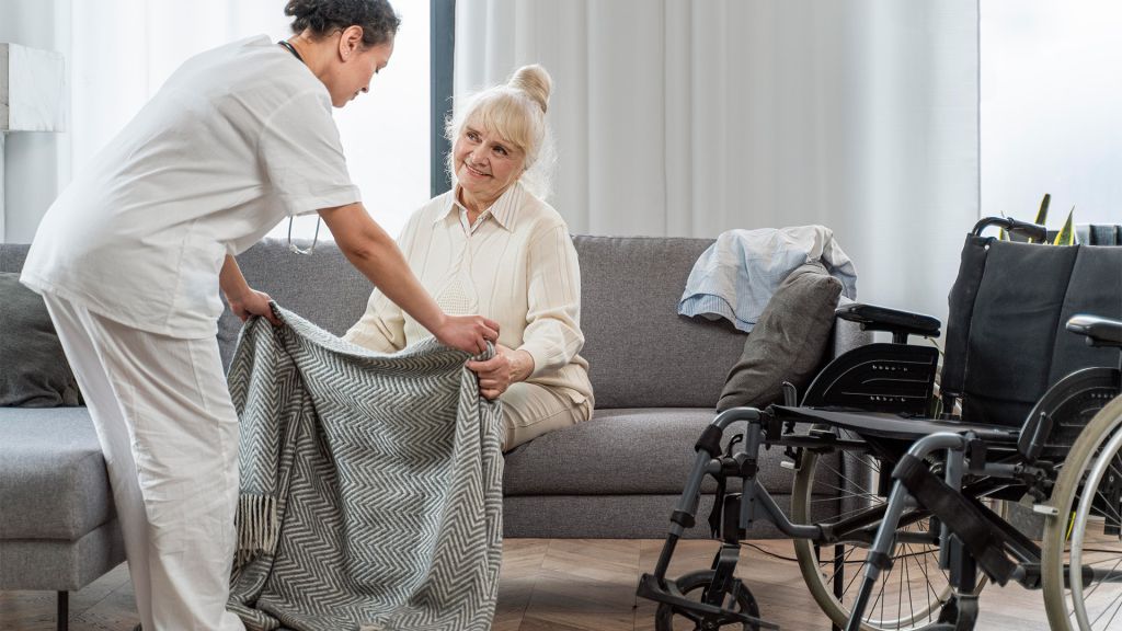 Pflegekraft deckt eine ältere Frau auf dem Sofa mit einer Wolldecke zu