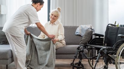 Pflegekraft deckt eine ältere Frau auf dem Sofa mit einer Wolldecke zu